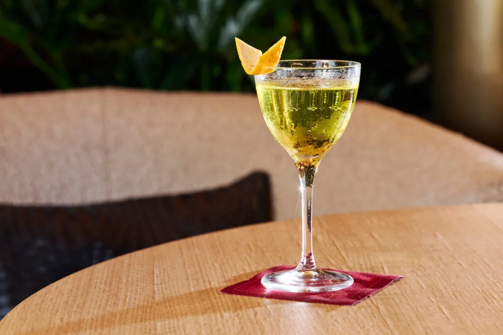 A cocktail in a stemmed glass with a citrus twist garnish sits on a red napkin on a wooden table, with a blurred background.