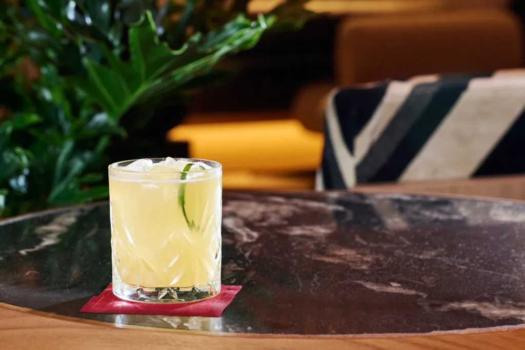 A glass of yellow cocktail with ice and a lime garnish sits on a red coaster on a dark marble table, with plants and soft seating in the background.