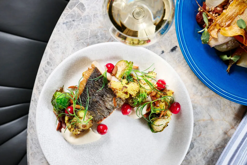 A plated fish dish garnished with Brussels sprouts, red berries, and herbs, served beside a glass of white wine on a marble table.