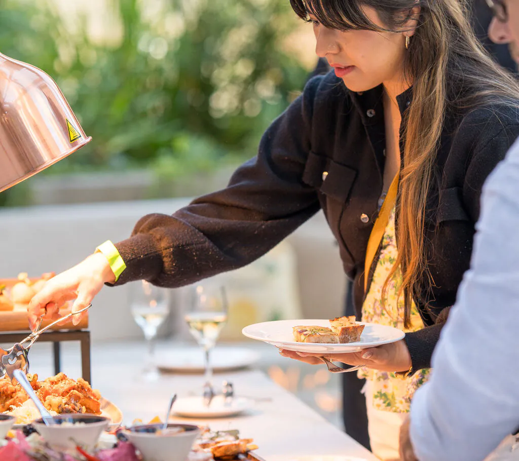 A woman serves herself food from a buffet table outdoors, holding a plate, with other people and dishes visible nearby.