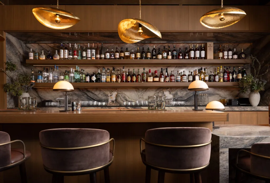 A stylish bar with velvet stools, golden pendant lights, and shelves filled with various bottles of liquor against a marble wall.