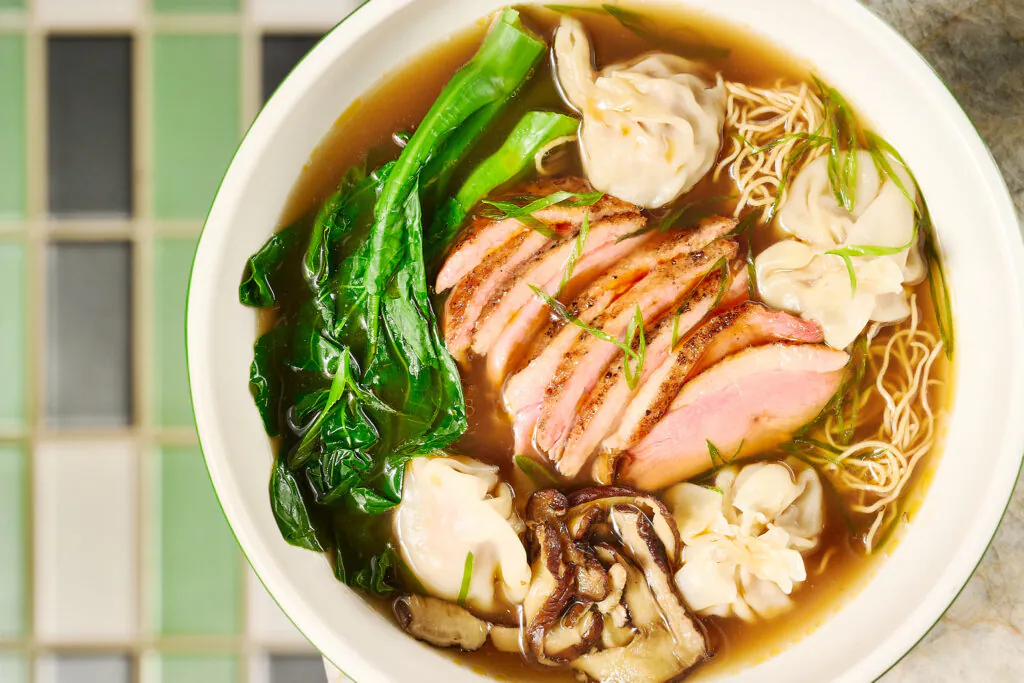 A bowl of noodle soup with sliced duck, dumplings, mushrooms, and leafy greens in broth, viewed from above on a tiled surface.