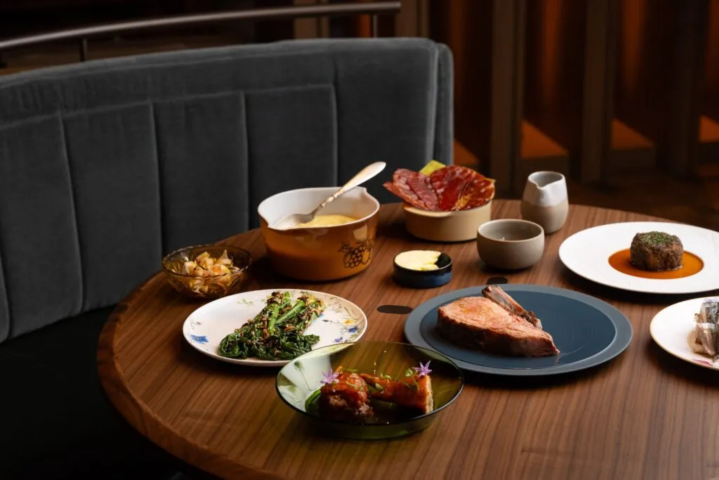 A wooden table set with plates of steak, vegetables, cured meats, sauces, and side dishes in a modern restaurant setting.