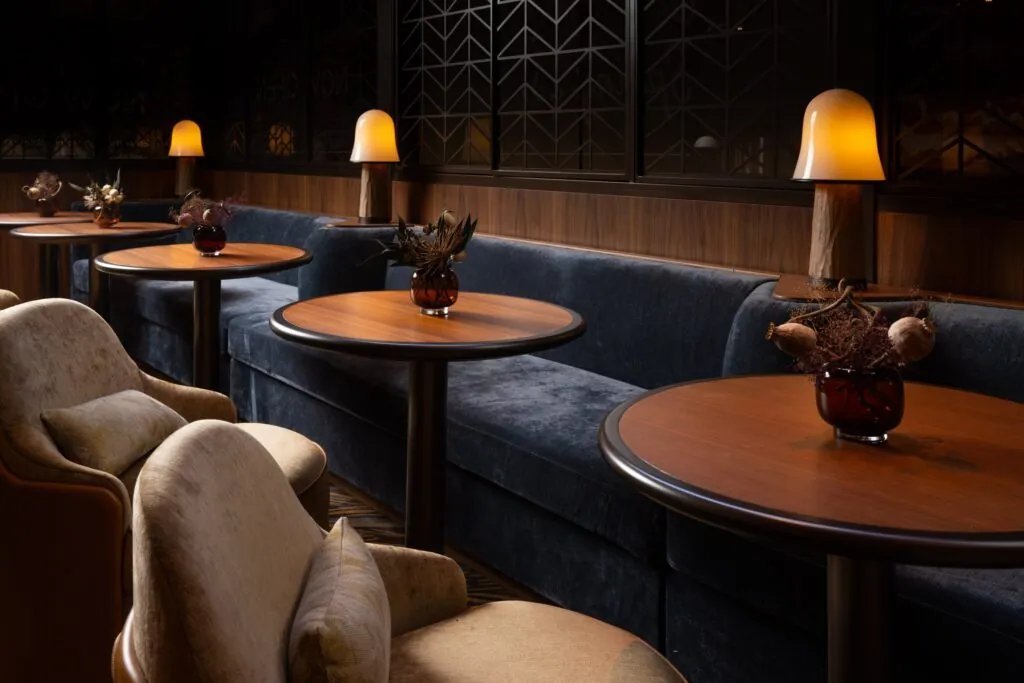 A cozy restaurant interior with round wooden tables, dark blue velvet seating, amber lamps, and dried floral centerpieces.