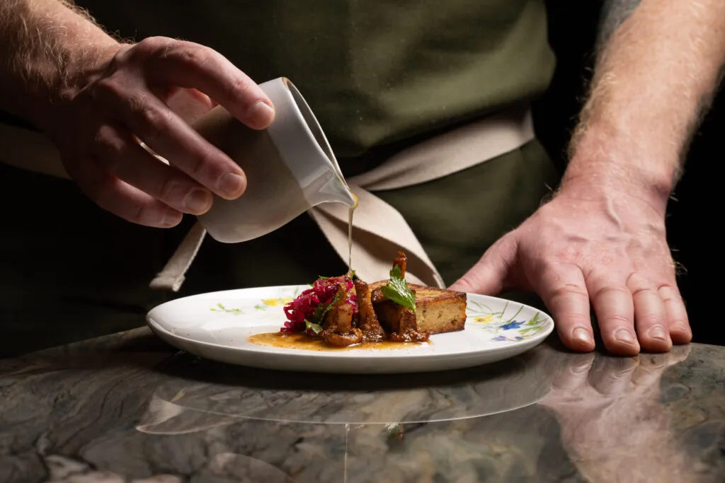 A person pours sauce from a small jug onto a plated gourmet dish with vegetables and garnishes on a marble surface.