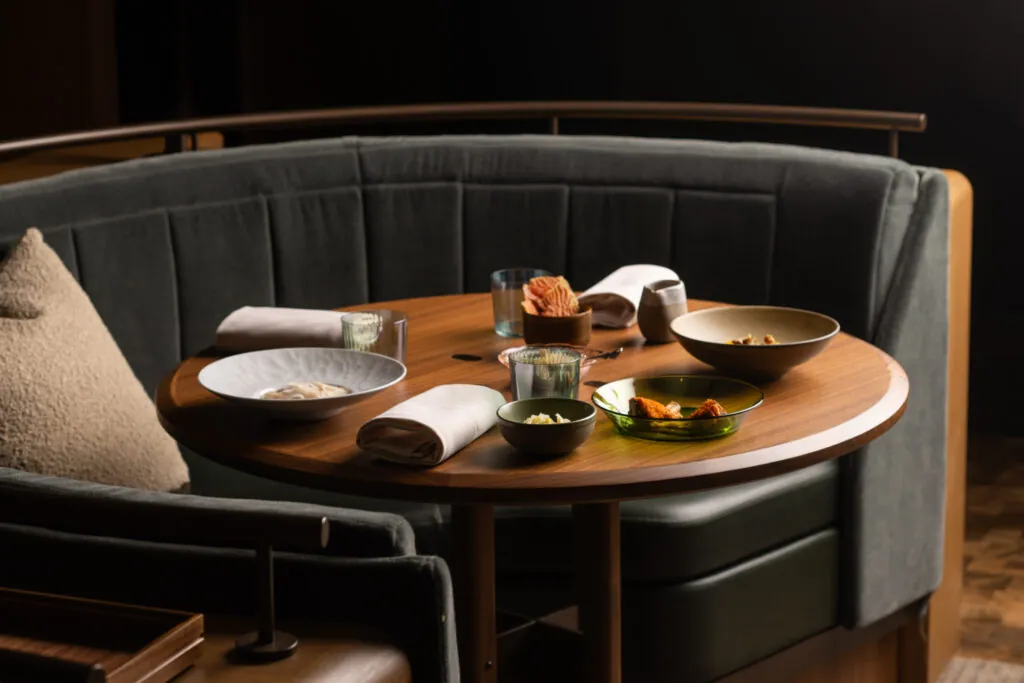 A round wooden table with food dishes, glasses, and napkins, set in a cozy, curved booth with gray upholstery.