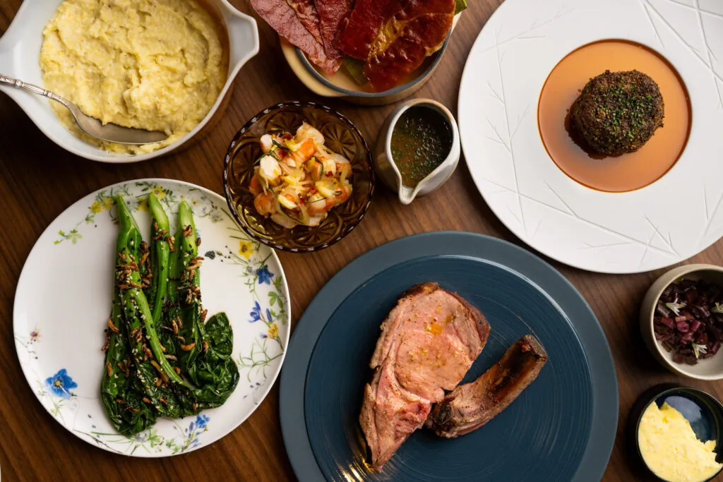 A table set with creamy polenta, glazed ham, a seasoned beef ball, sautéed greens, a grilled pork chop, and various sauces and condiments.