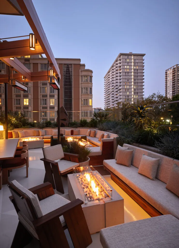Rooftop lounge at dusk with cushioned seating, a fire pit, and city buildings in the background under a clear evening sky.