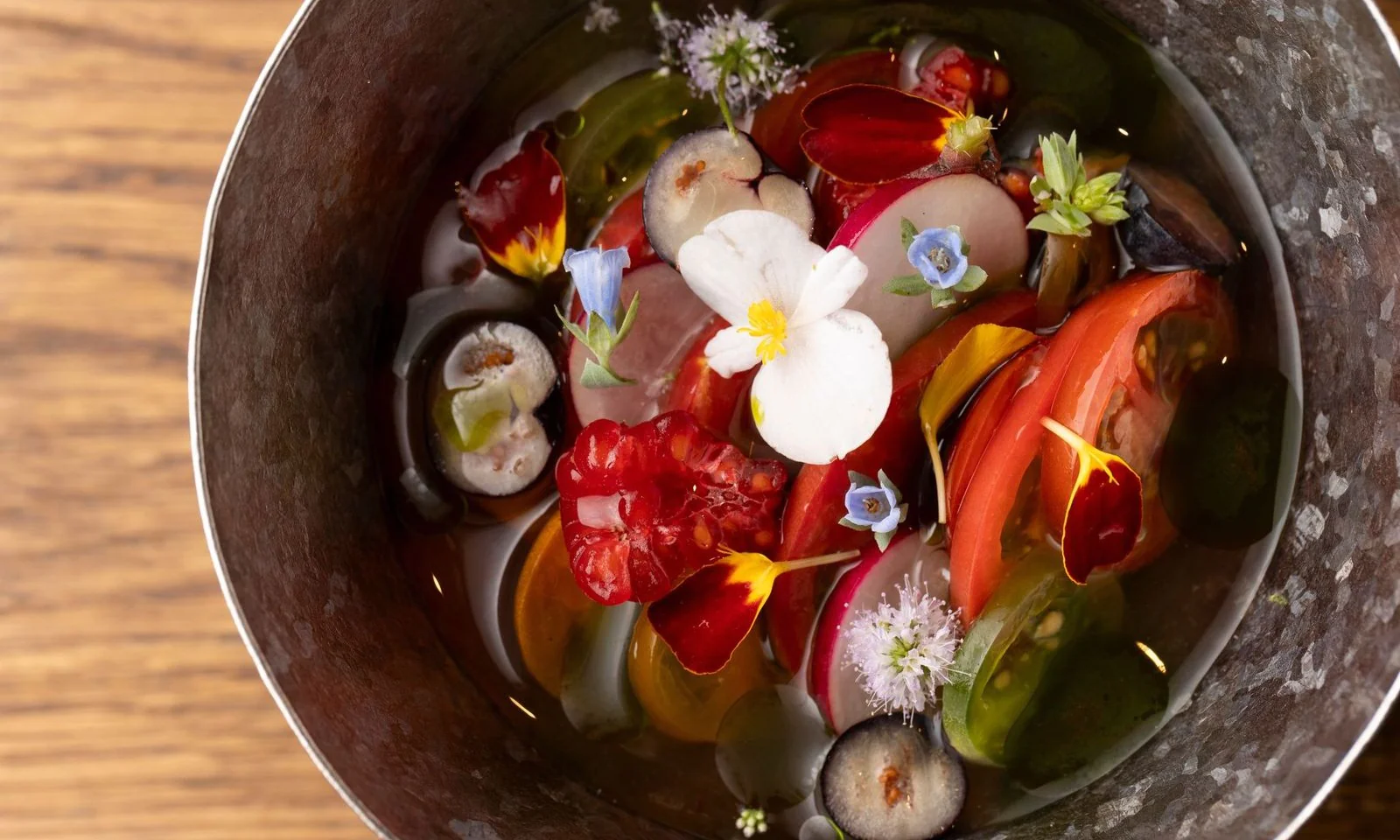 This bowl features a mix of sliced radishes, fresh tomatoes, berries, and various edible flowers, all served in a rustic metal container on a wooden surface.