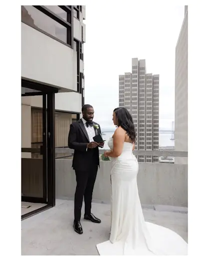 A bride and groom stand on a city rooftop, facing each other and exchanging vows, with tall buildings and a bridge in the background.
